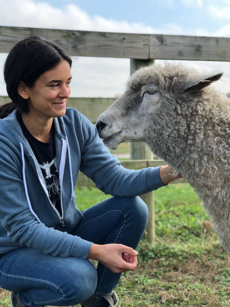 Image of the PAVFA Co-Founder and Board Secretary Jennifer Ramos-Buschmann, sitting on her heels in a grassy area inside a farm, petting a sheep.