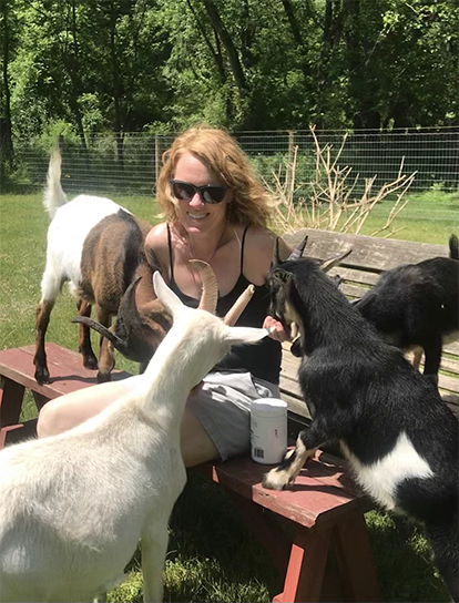 Image of Sheryl Petrillo, co-founder and board member of PAVFA, sitting on a red bench in a grassy farm area, attempting to feed four goats with her hands.